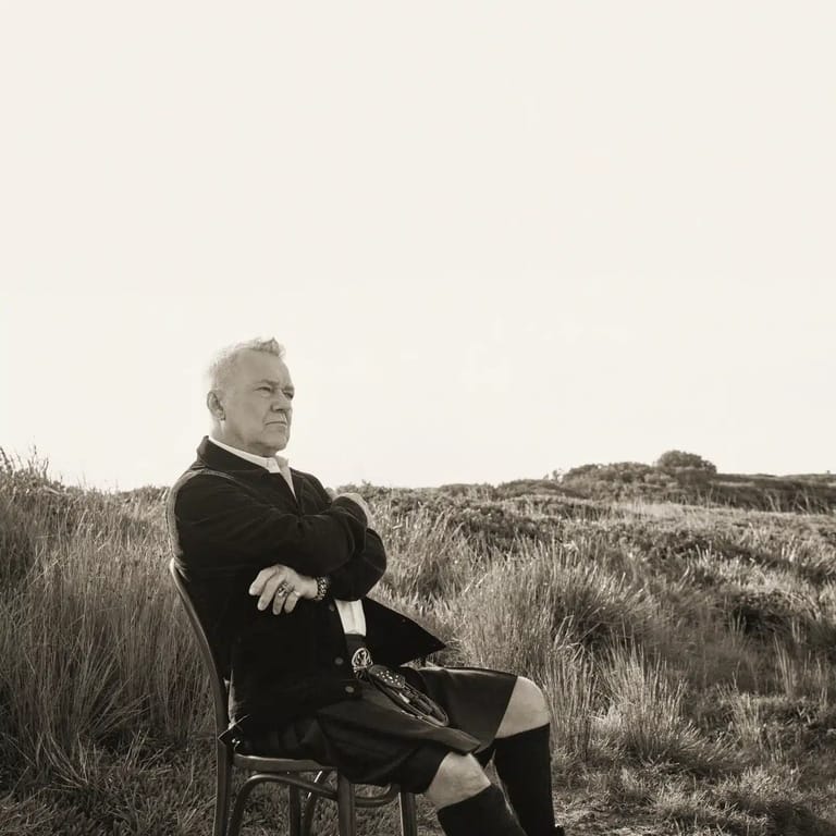 Man in dark jacket seated on chair in sparse, dry grassland under overcast sky.