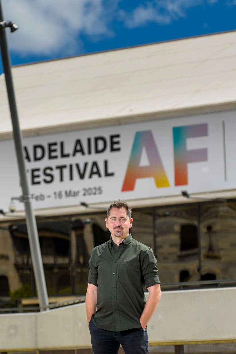 Man in dark shirt standing before Adelaide Festival signage under blue sky.