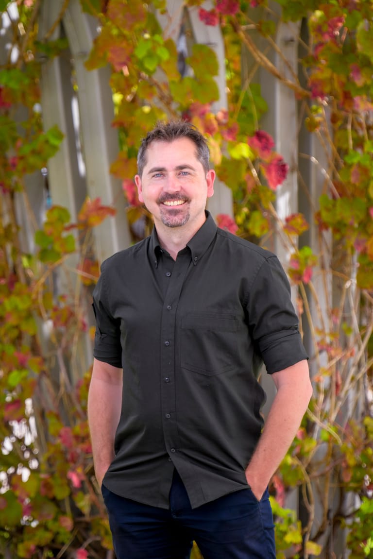 Man in black shirt smiling against a backdrop of autumn ivy and climbing vines.
