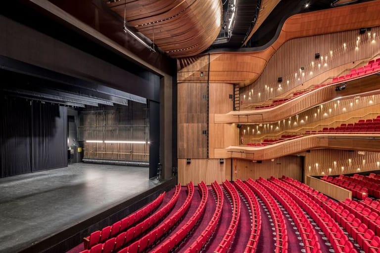 Empty theatre auditorium with rows of red seats facing a dark stage framed by warm wooden architecture.