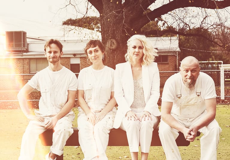 Four people in white clothing seated together on grass beneath a large tree in warm afternoon sunlight.