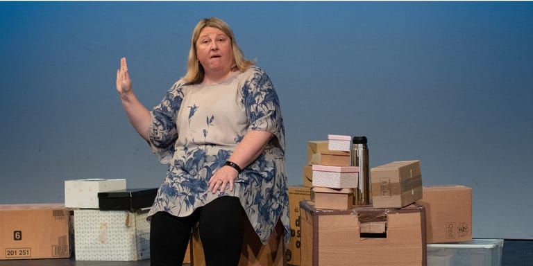 Performer gestures expressively surrounded by cardboard boxes against a blue backdrop during a theatre production.