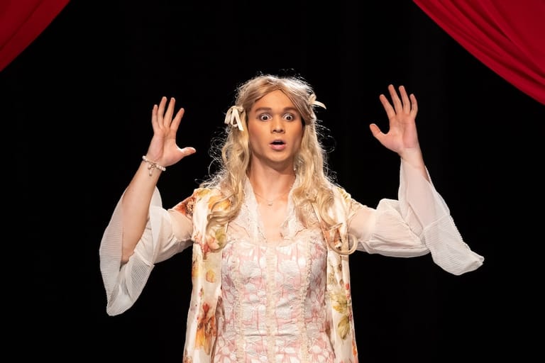 Performer with long blonde hair in period costume with hands raised expressively against red stage curtains.