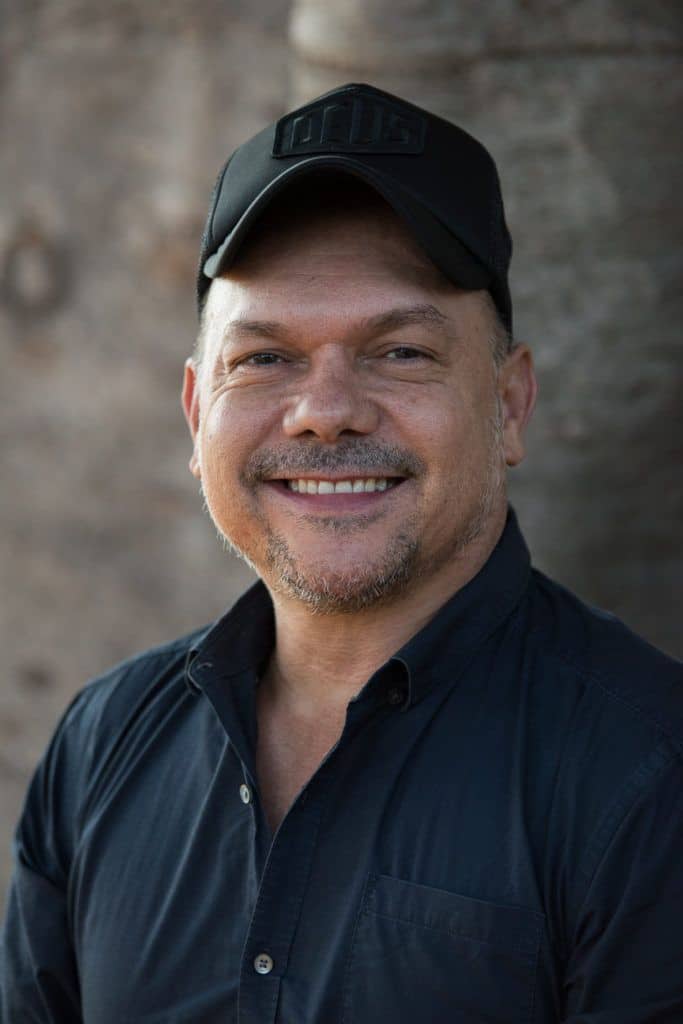 Man in black cap and shirt smiling at camera against neutral textured background.
