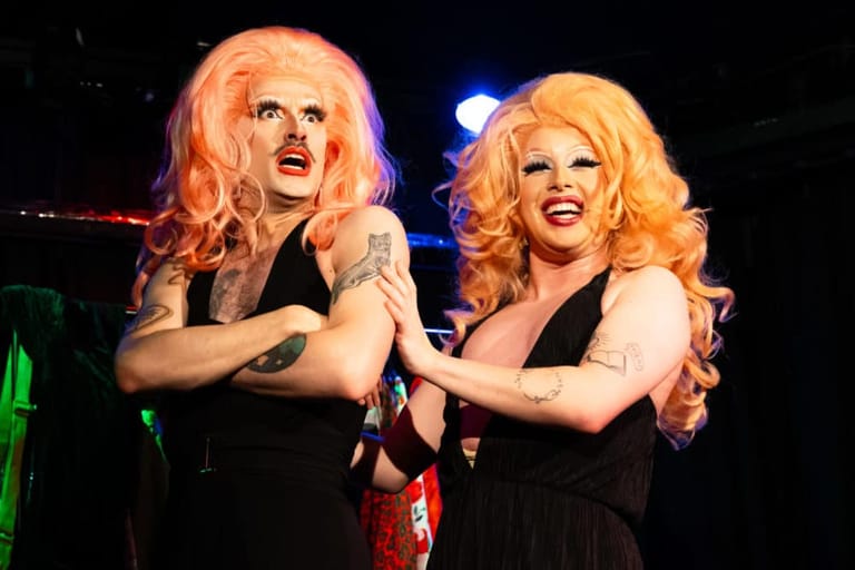 Two performers with blonde wigs in black outfits perform together on stage under coloured lights.