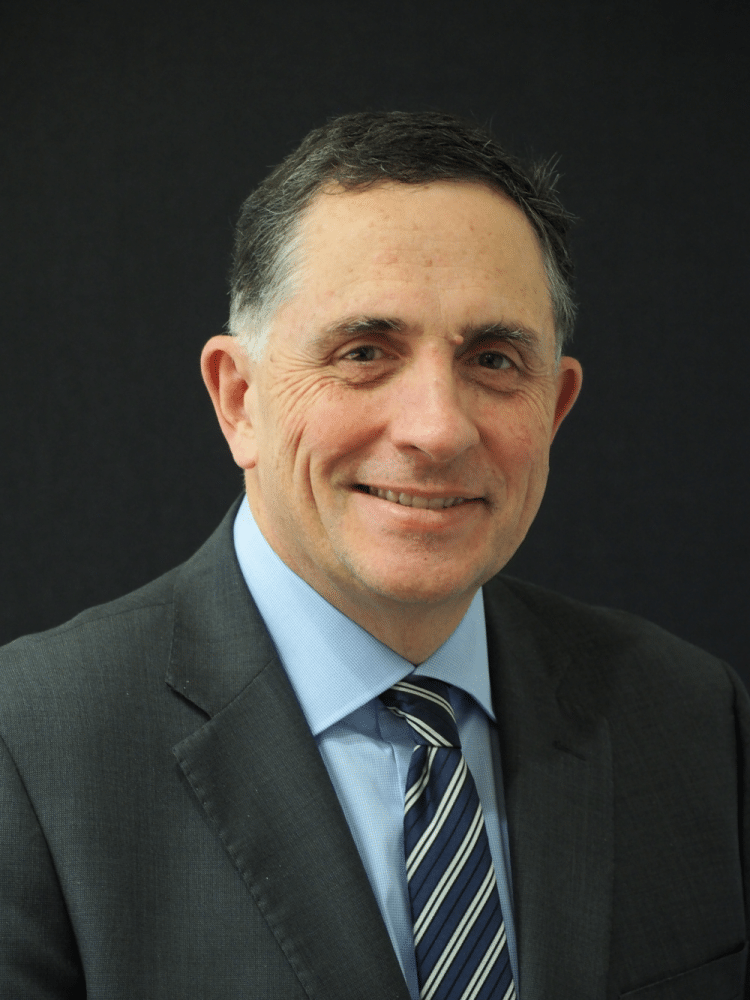 Man in dark suit and light blue shirt with striped tie, smiling against a dark background.