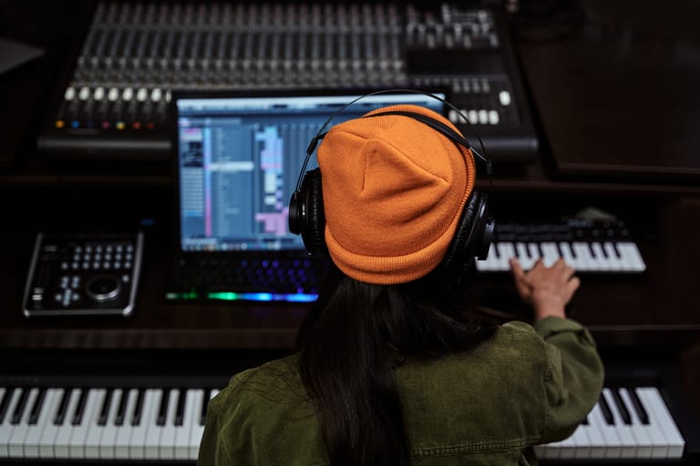 Music producer wearing orange beanie and headphones works at mixing console with keyboards and monitors.
