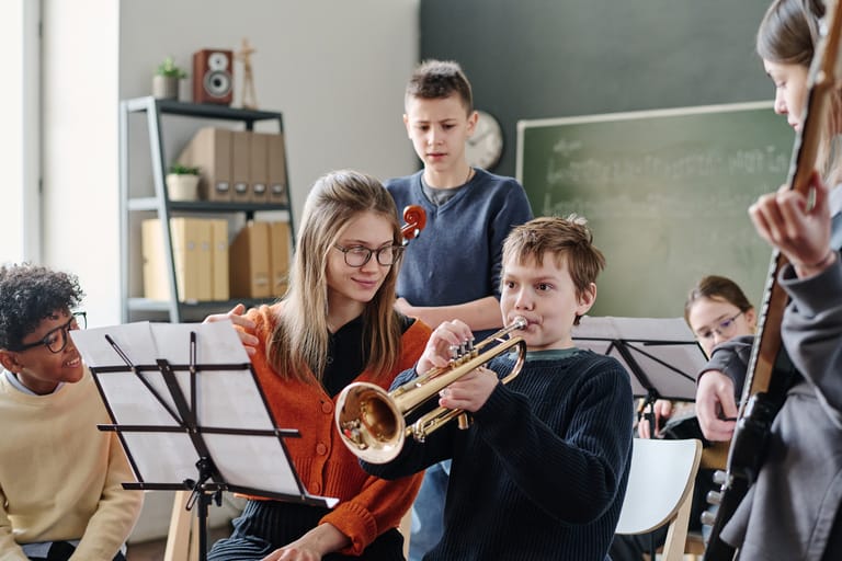 Young musicians play brass instruments during a classroom music lesson with sheet music stands and a blackboard visible.