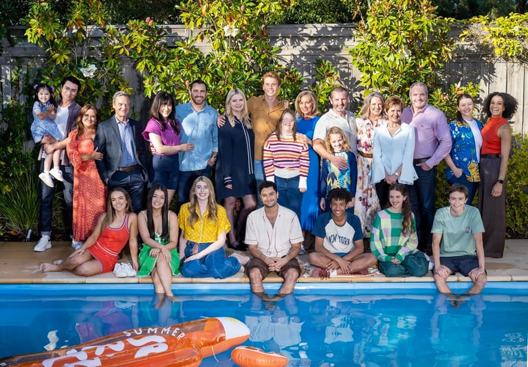 Large group of people of varying ages posed together by a swimming pool with green foliage behind them.