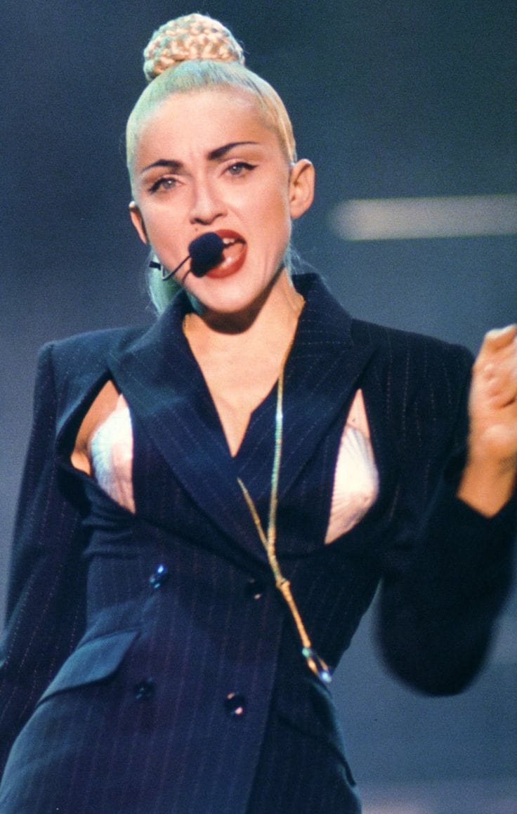 Performer singing into a headset microphone wearing a dark blazer and blonde hair styled in a top knot.