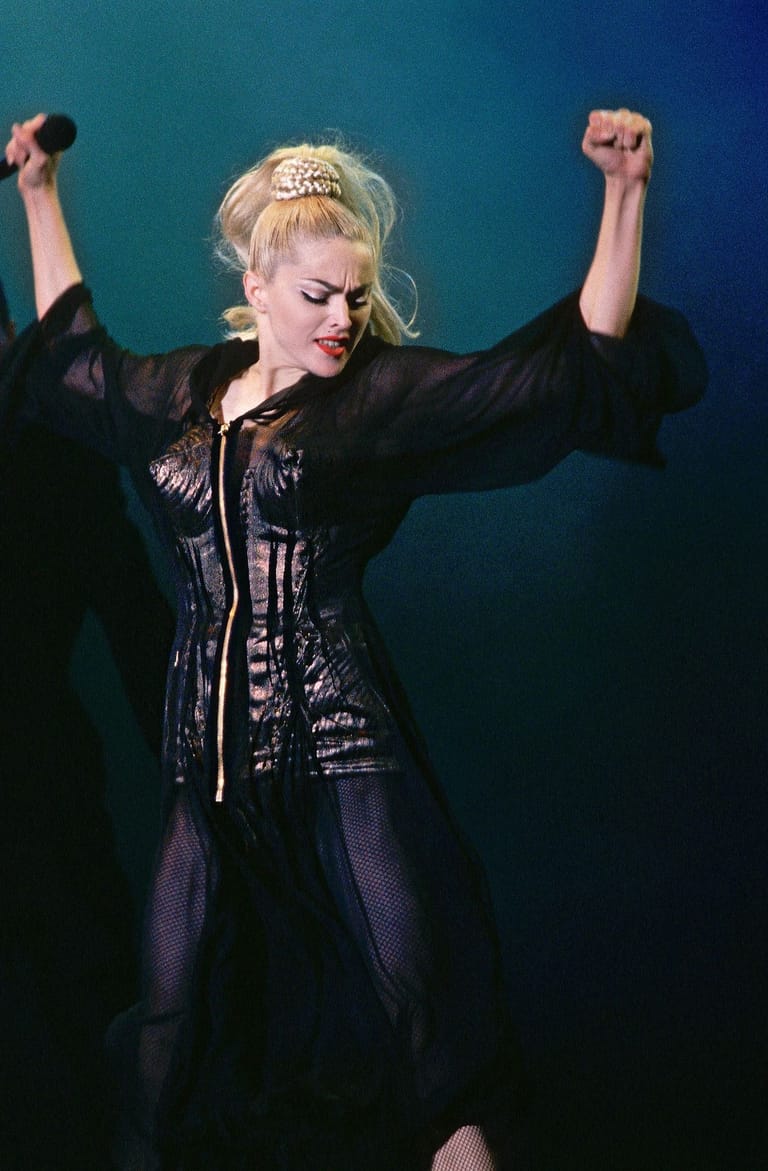 Performer in black sequined jacket and blonde hair raises fists triumphantly against teal and blue stage lighting.