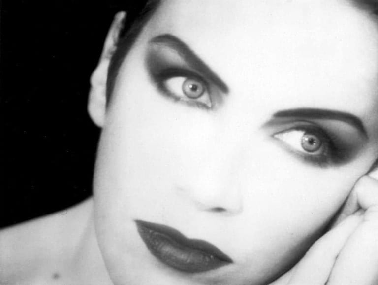 Close-up portrait of a person with dramatic black and white makeup and dark lipstick against a black background.