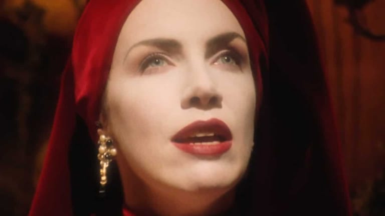Person with red hair and lipstick wearing gold earrings, lit by warm red stage lighting.