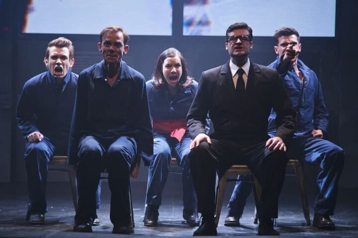 Five performers seated in a row on stage, illuminated by blue lighting against a dark backdrop.