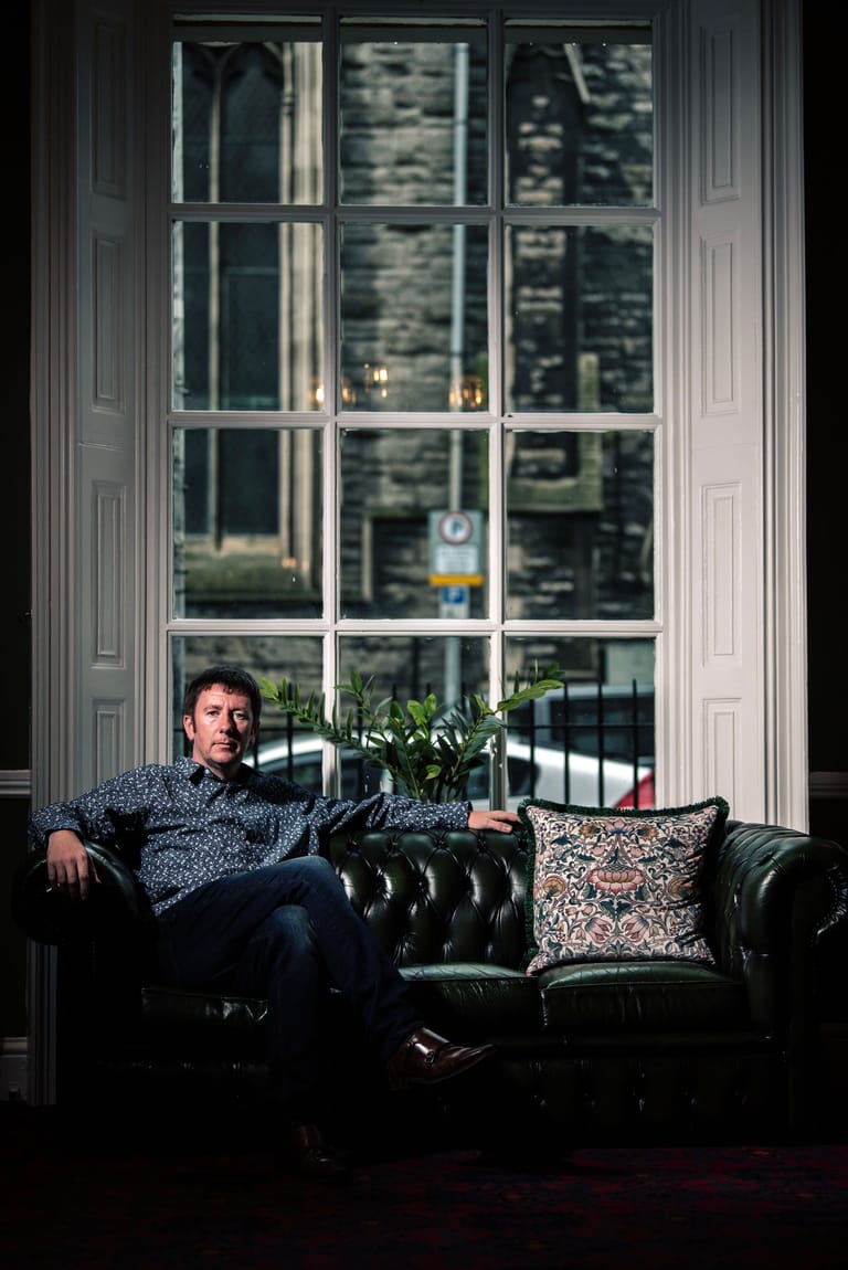 Man seated on a black leather Chesterfield sofa against a tall sash window in an elegant interior space.