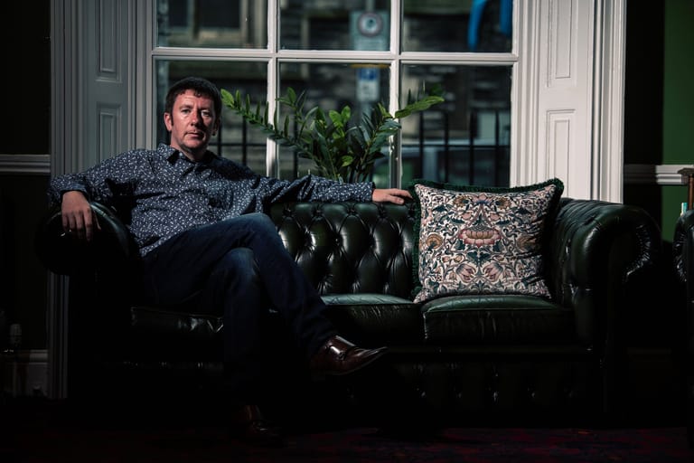 Man in patterned shirt reclines on black leather sofa against bright windows, relaxed tour portrait setting.