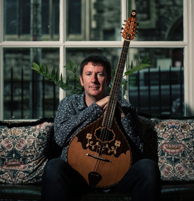 Musician holding a wooden acoustic guitar seated on a patterned couch against large windows.