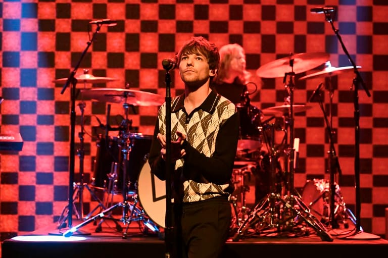 Musician in geometric patterned jacket performing with acoustic guitar under red stage lighting, drummer visible behind.
