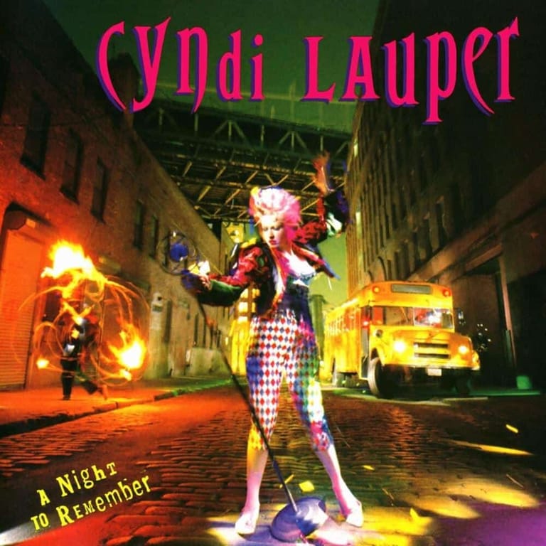 Performer in vibrant clown costume with pink hair stands on colourful urban street set with vintage yellow cars and neon lighting.