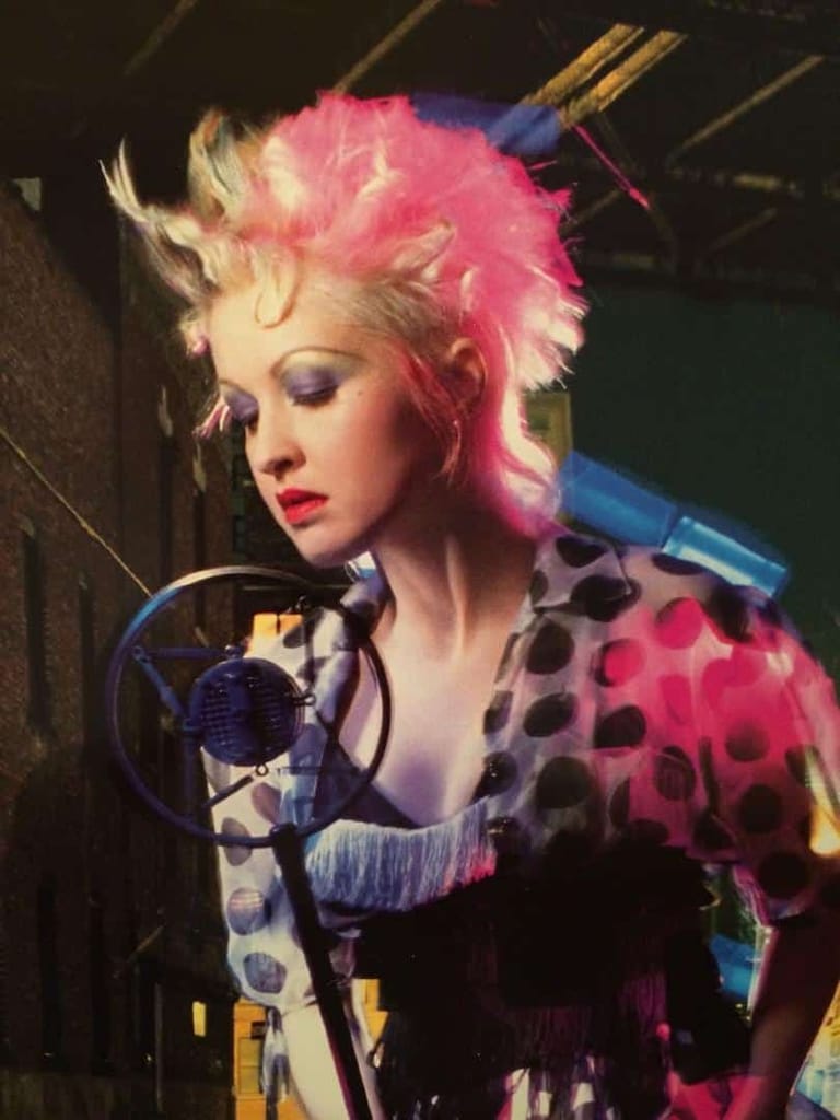 Singer with voluminous pink hair performing at microphone under stage lighting.