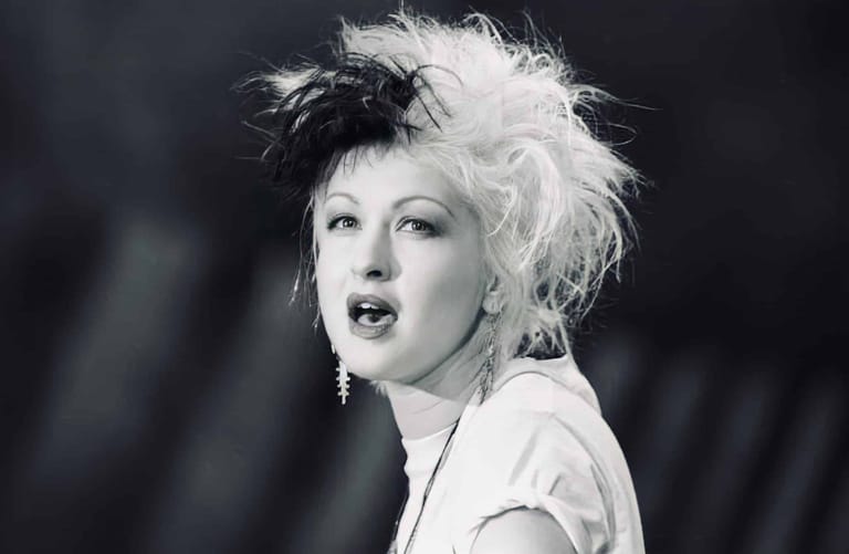 Person with voluminous black and white two-toned hair and pale lipstick against a dark background.