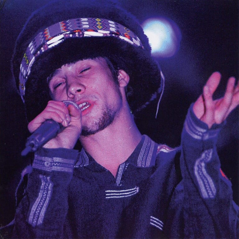 Performer singing into microphone wearing a striped shirt and patterned headband, gesturing expressively under stage lighting.