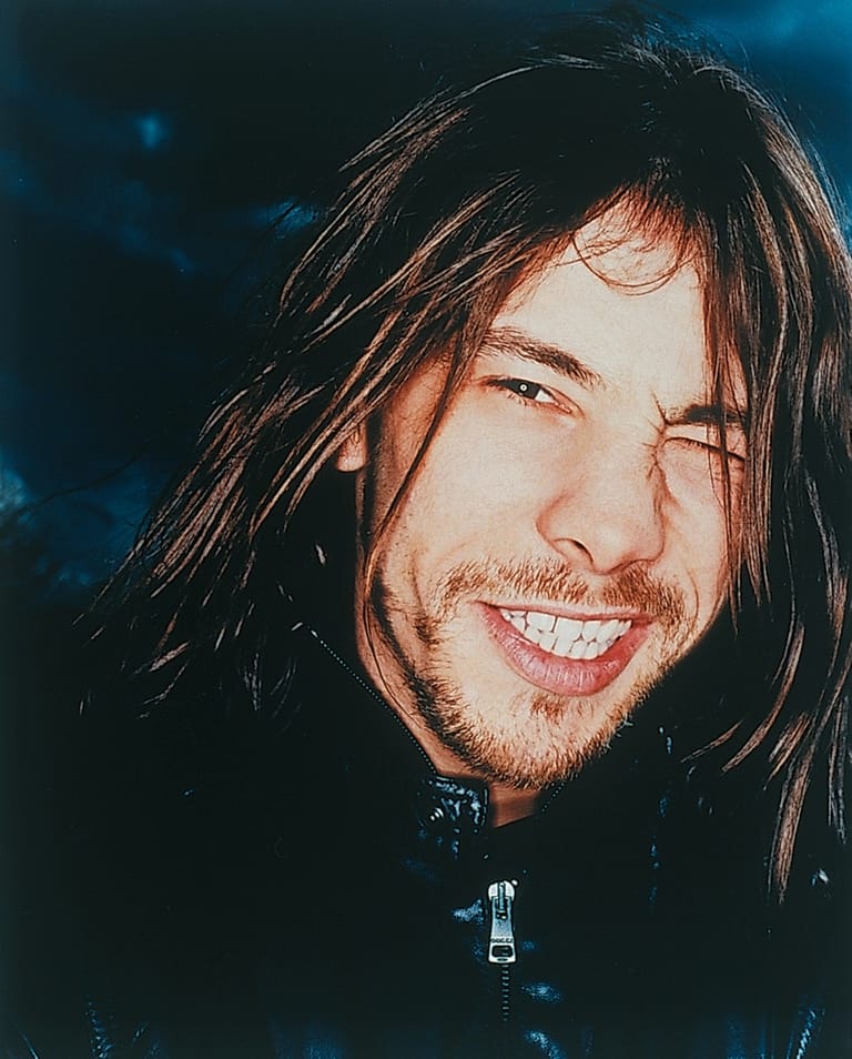 Man with long dark hair smiling against a dark blue background.
