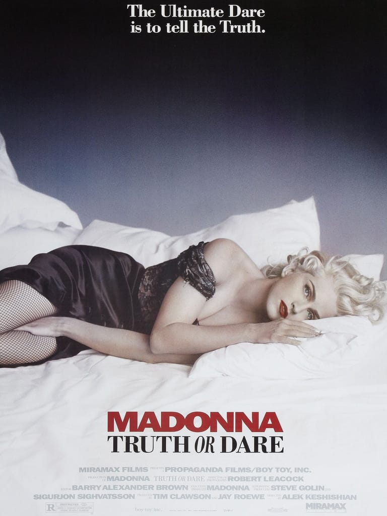 Person in black lingerie reclining on white bedding with platinum blonde hair against moody blue-grey background, promotional movie poster.