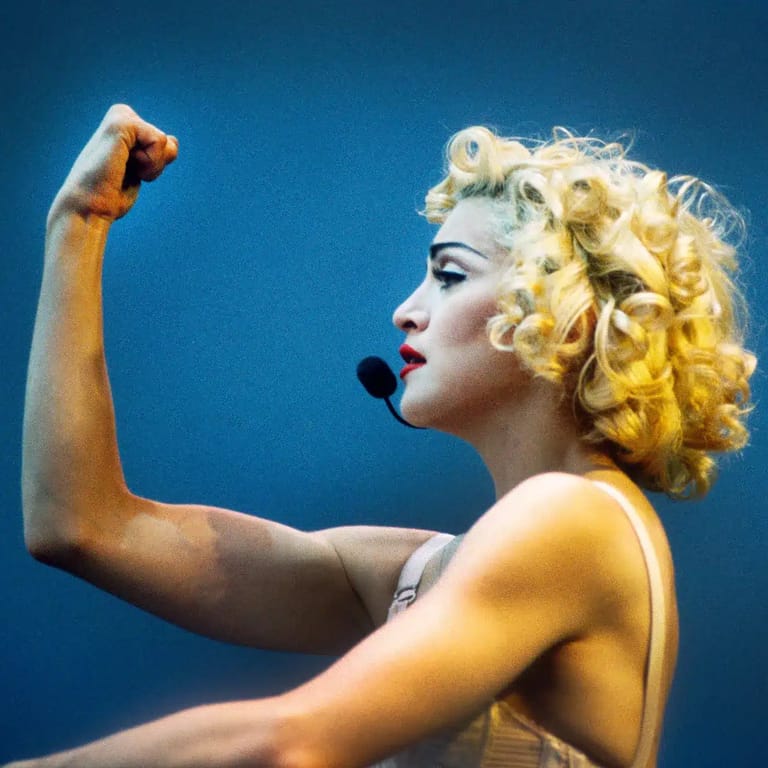 Performer with blonde curled hair flexing her arm in a power pose against a deep blue background.