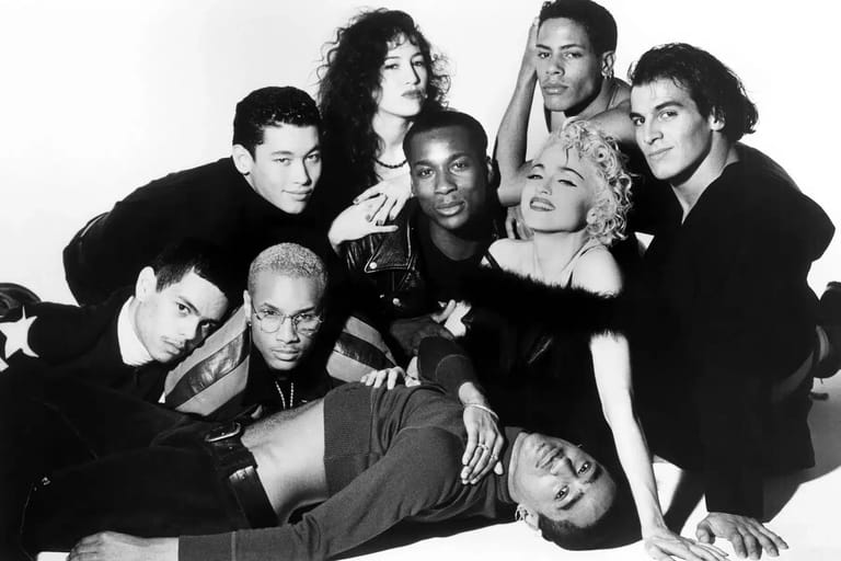 Group of nine people in black and white studio portrait, arranged closely together in playful poses.
