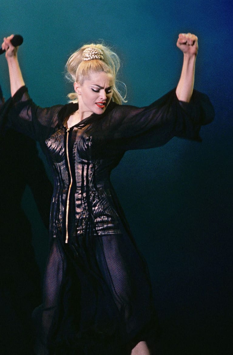Blonde performer in black sequined jacket and headband raises both fists triumphantly against a teal-blue stage backdrop.