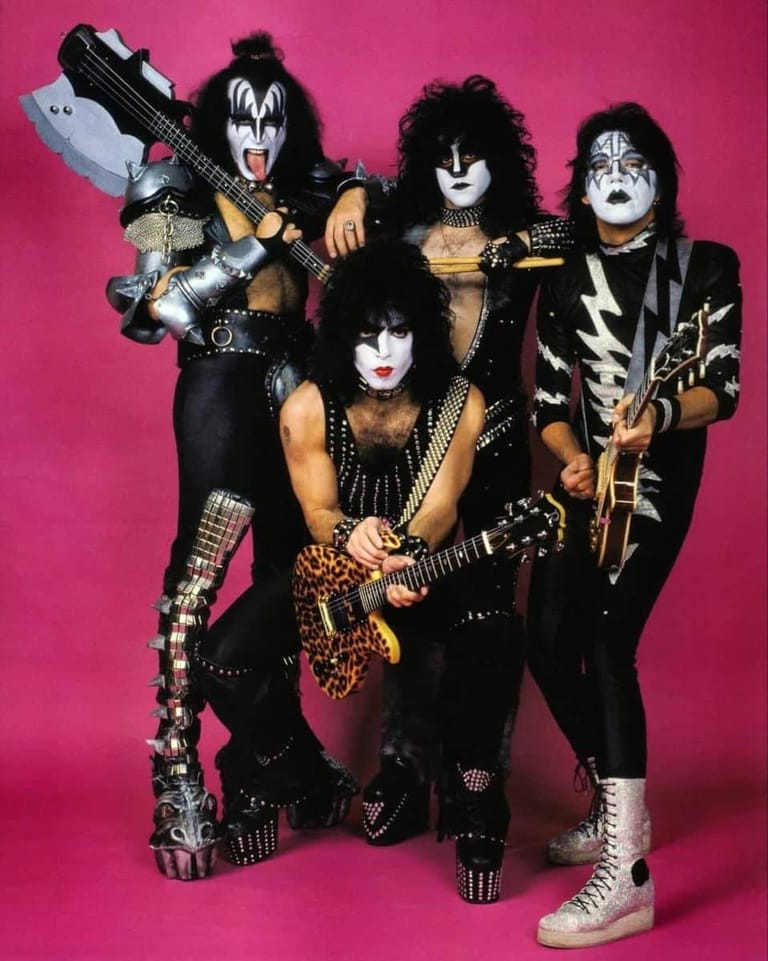 Four musicians in theatrical face paint and studded black leather costumes pose against a hot pink background with electric guitars.