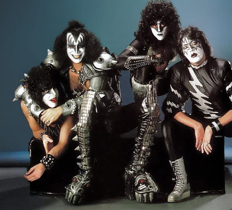 Four musicians in full theatrical makeup and studded black leather costumes pose against a blue backdrop.