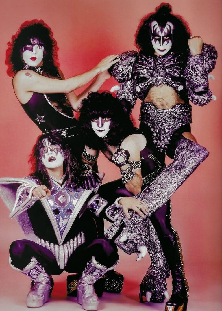 Four musicians in full theatrical makeup and glam rock costumes pose together against a pink backdrop.