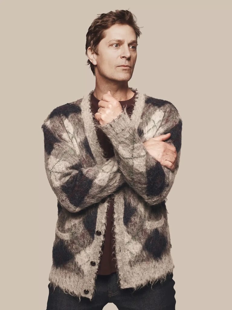 Man in a patterned knit cardigan with arms crossed, gazing to the side against a neutral background.