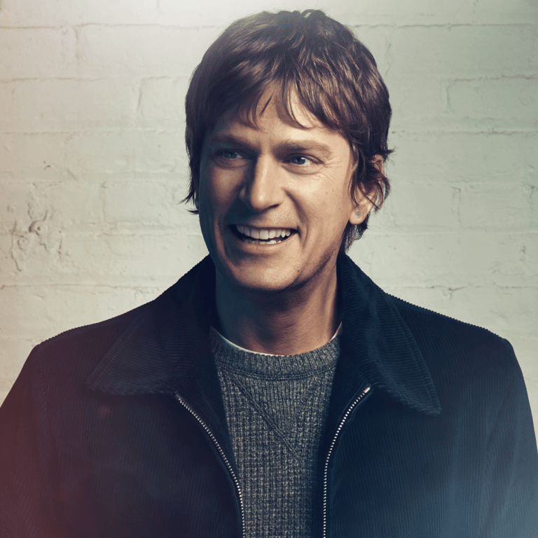 Man with dark hair smiling at camera against white brick wall, wearing black jacket over grey sweater.