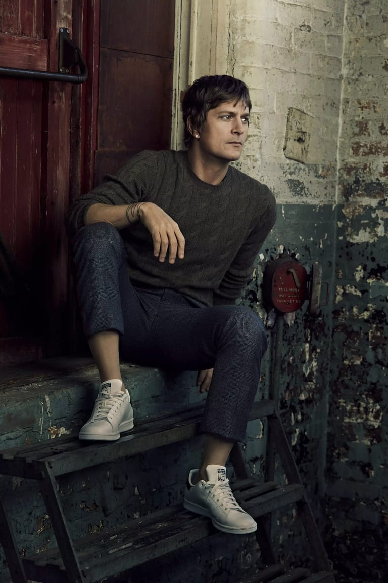 Man in dark shirt and jeans seated on weathered metal steps against peeling painted wall.