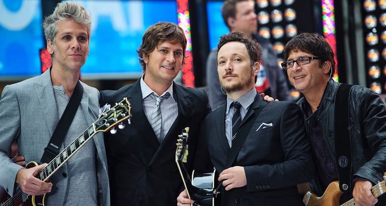 Four musicians stand together on a brightly lit stage holding guitars and awards.