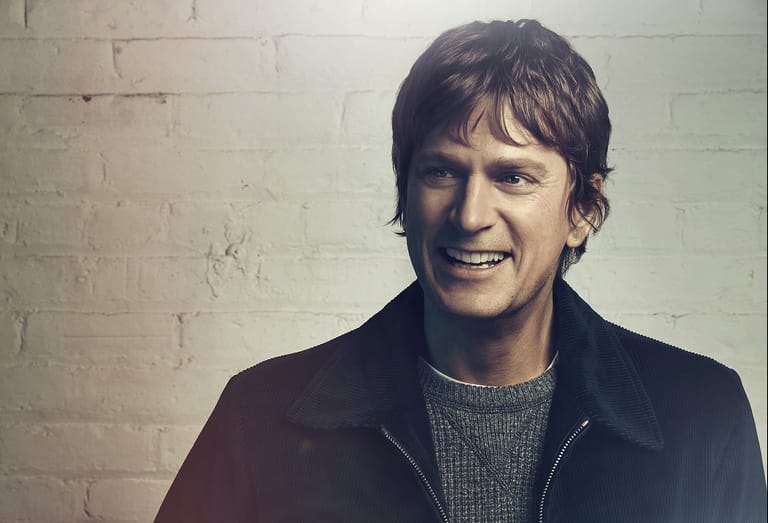 Man with dark hair smiling at camera against a white brick wall.