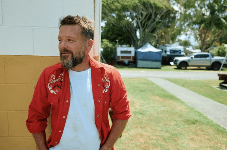 Man in red embroidered jacket stands outdoors against a cream-coloured wall, smiling.