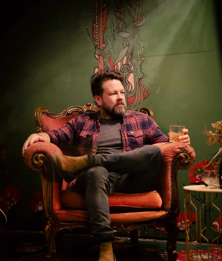 Bearded musician seated in ornate armchair on stage, lit by warm amber spotlight against artistic backdrop.