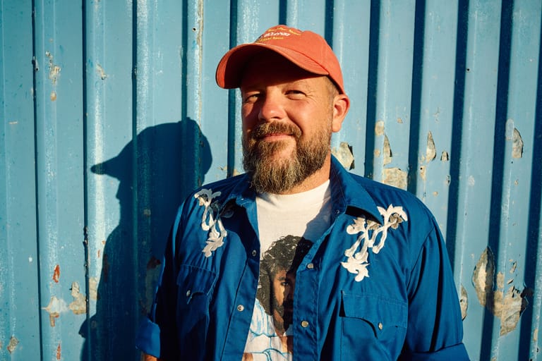 Bearded man in orange cap and blue jacket against weathered corrugated metal wall.