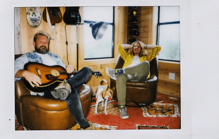 Bearded musician holding an acoustic guitar sits opposite a woman in a studio space cluttered with instruments and artwork.