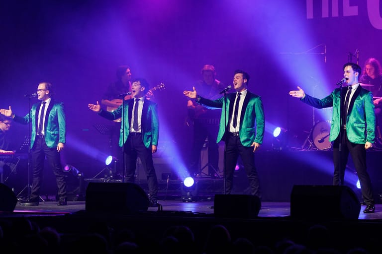 Five performers in matching teal and navy suits strike synchronized poses under purple stage lighting during a concert performance.