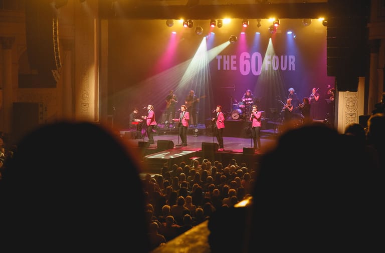 The Go Four band performs on stage under coloured spotlights with audience silhouettes in the foreground.