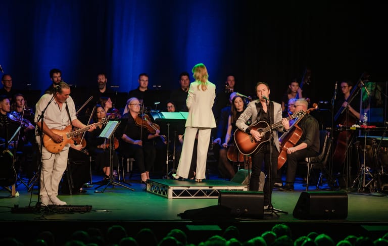 Large ensemble of musicians and a central singer in cream suit performing on stage under blue lighting.