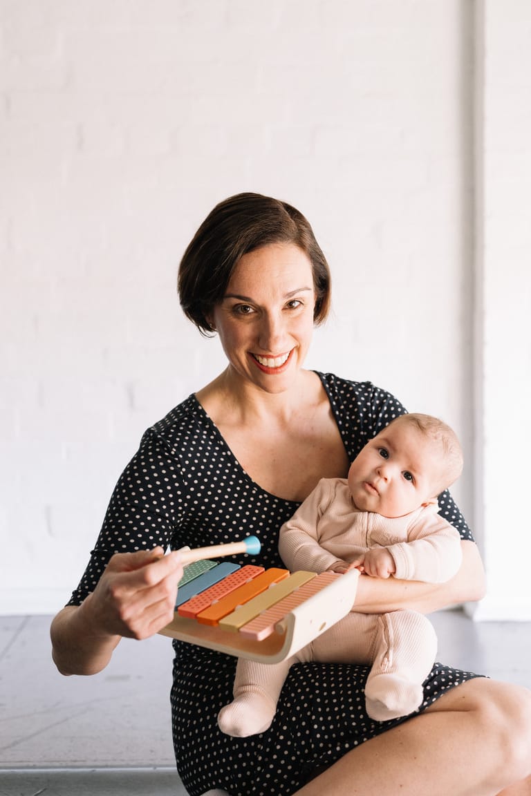 Woman in polka-dot dress holding infant while displaying a colourful wooden toy against a white background.