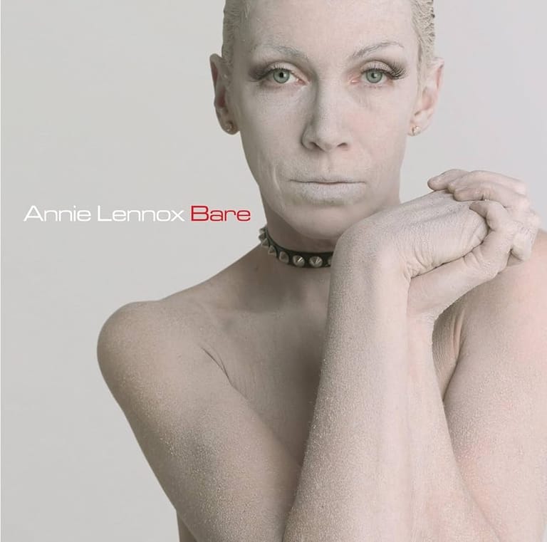 Bald person with choker necklace resting chin on clasped hands against neutral background.