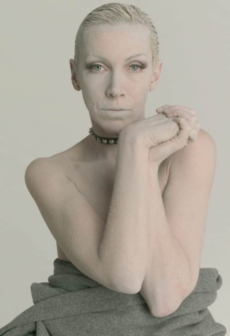 Person with slicked-back blonde hair and choker necklace poses with clasped hands against neutral background.