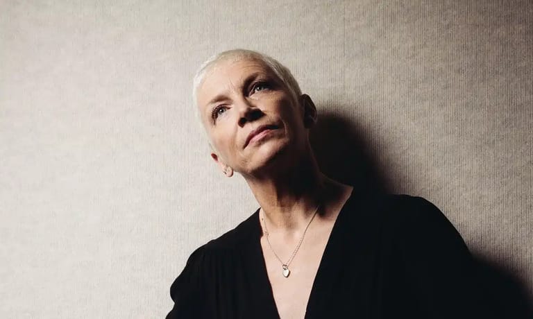 Person with short blonde hair wearing black jacket gazes upward against neutral background.